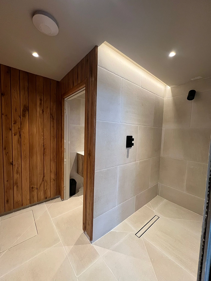 Modern bathroom with wooden wall, tiled shower area featuring black fixtures and linear drain, recessed ceiling lights, and doorway leading to sink
