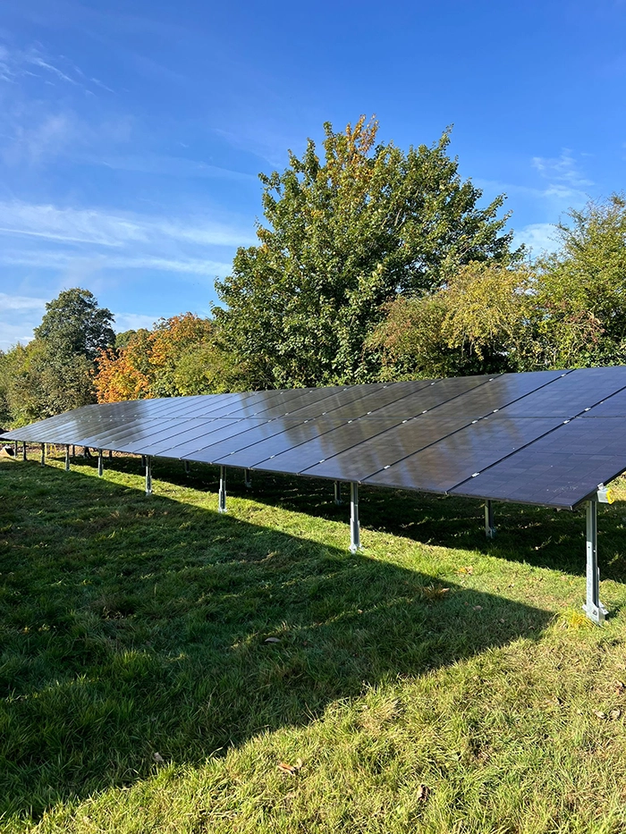 Row of solar panels mounted on metal frames in grassy field, angled toward sun with trees and clear blue sky in background
