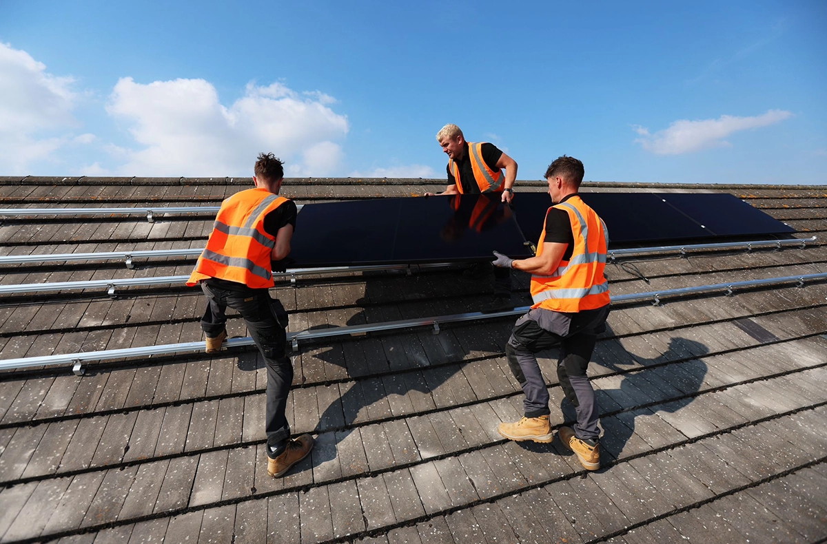 Three workers in orange high-visibility vests installing solar panels on sloped roof under clear sky
